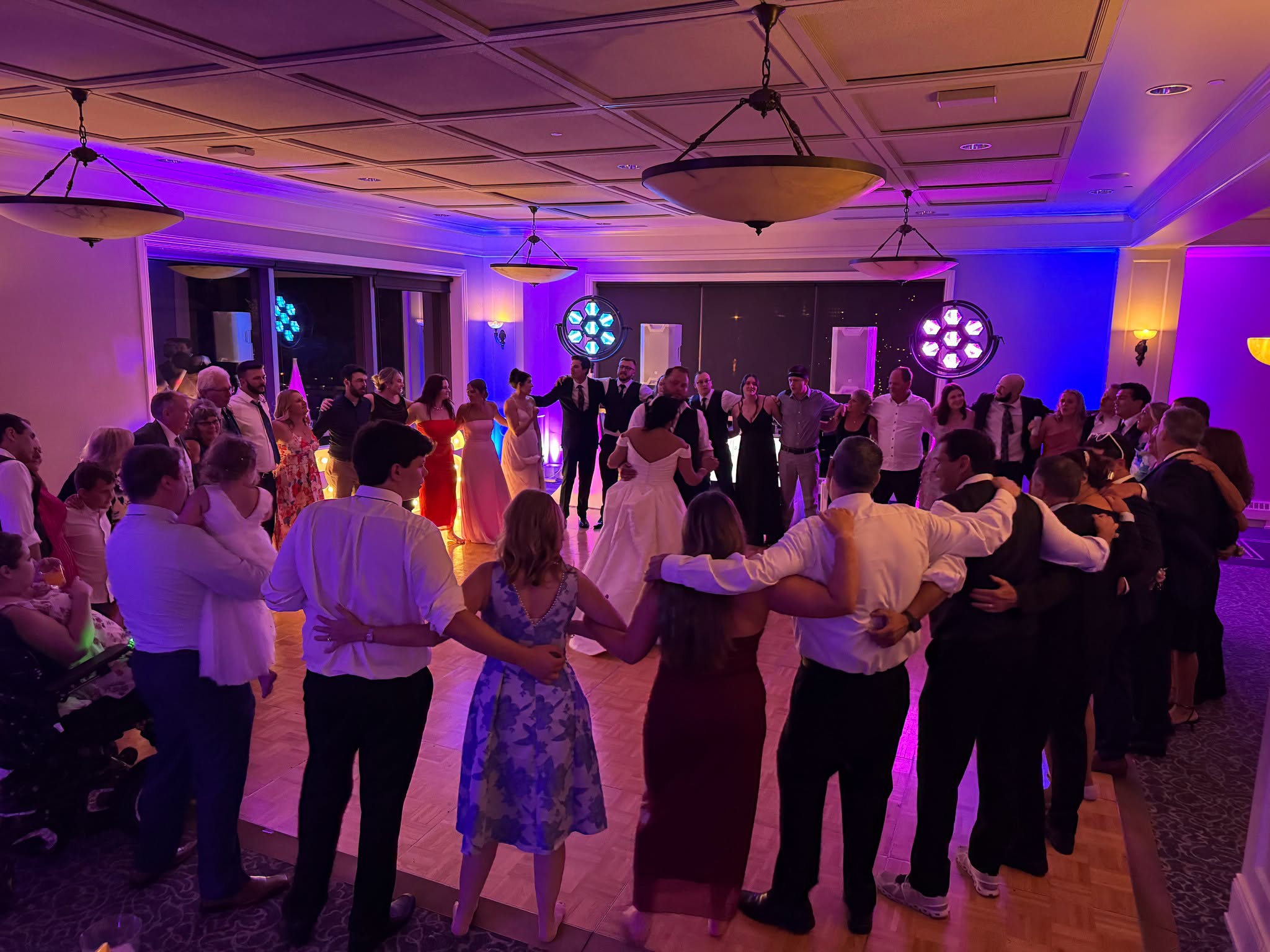 Wedding guests forming a circle with their arms around each other around the newlyweds under purple lighting